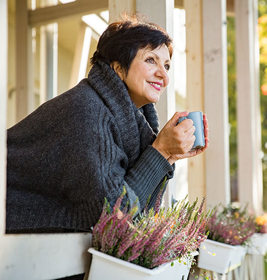 Mature attractive woman standing on Cozy wooden terrace with cup of hot coffee wrapped up in knitted warm sweater, happy smile. Lake house in autumn, yellow and red leaves on trees. Sunny day