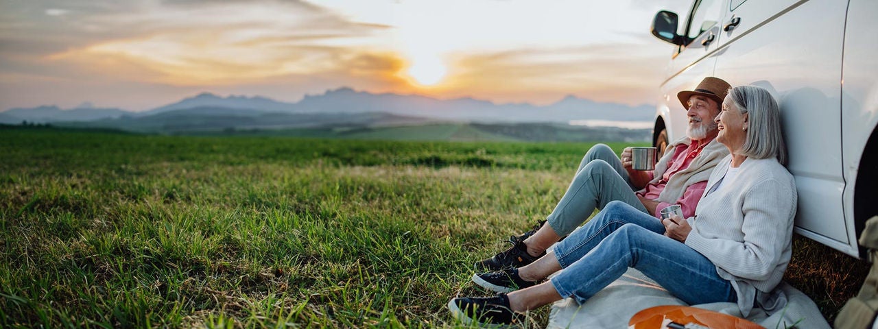 Senior couple sitting by car and drinking coffee after long drive during their roadtrip. Elderly spouses at autumn roadtrip watching the sun set behind High Tatras.