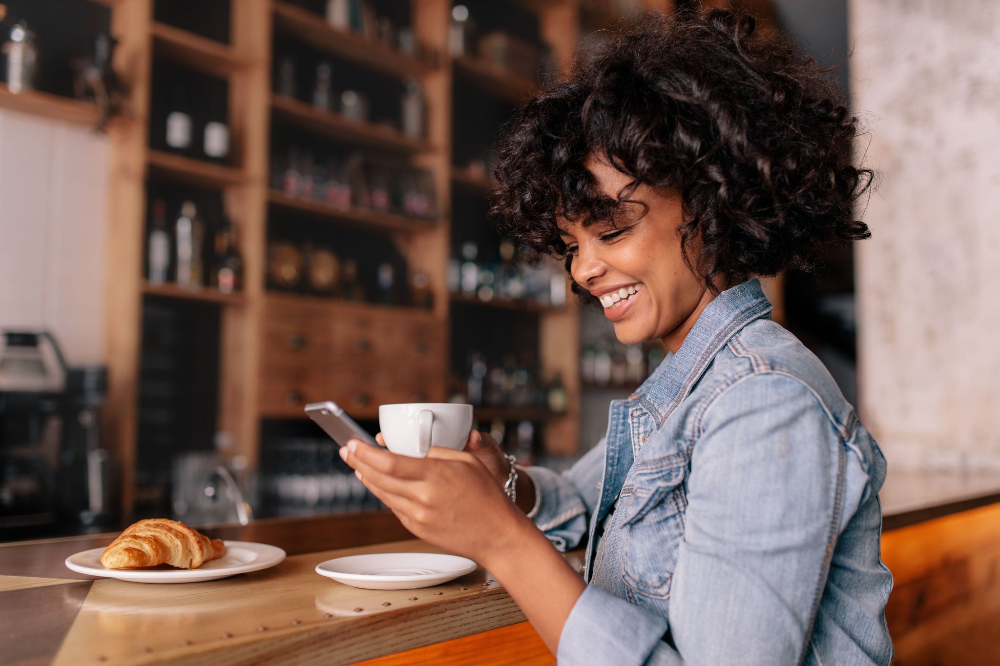 Smiling woman using smart phone in a modern cafe. Young african female sitting at a cafe counter having coffee and reading text message on mobile phone.