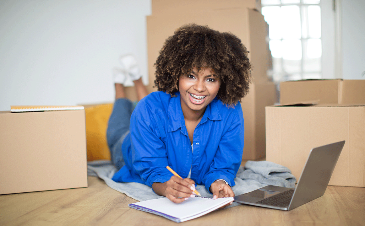 Cheerful Black Lady Making Checklist And Using Laptop While Moving Home