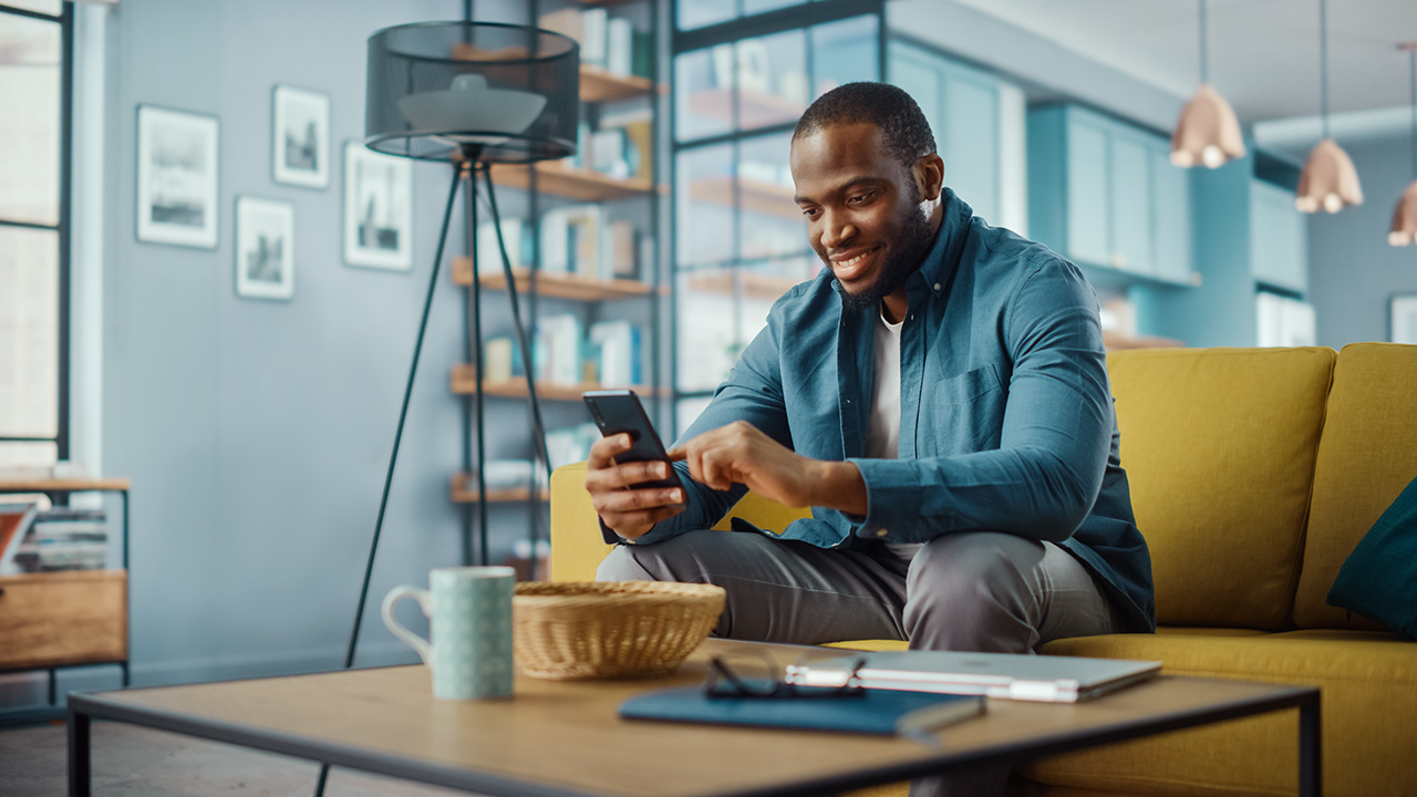 Handsome Black African American Man Using Smartphone while Sitting on a Sofa in Cozy Living Room. Freelancer Working From Home. Browsing Internet, Using Social Networks, Having Fun in Flat.